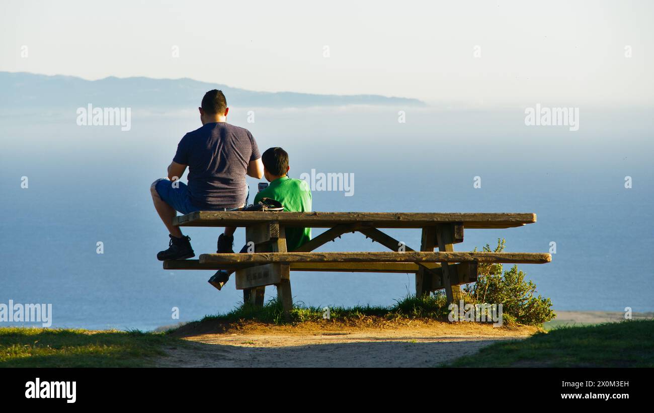 Two hikers rest on a picnic table facing the pacific ocean, enjoy the view. Stock Photo