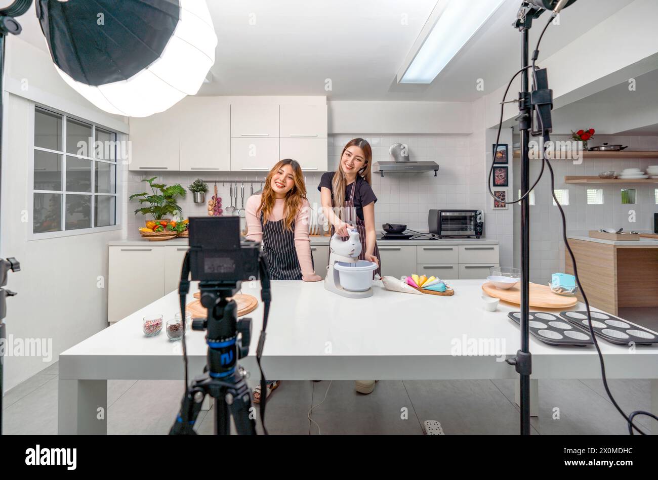 Two asian women in an apron, smiling and cooking in a kitchen with camera and lighting equipment for video and photo shoot, recording vlog video for s Stock Photo