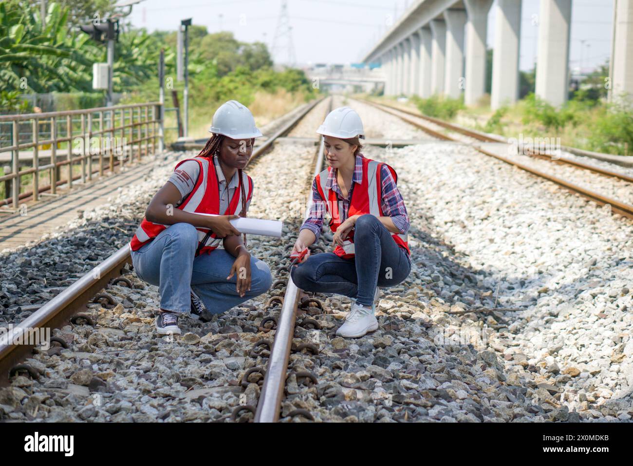 Rail transportation engineer in safety vest and hardhat check the neatness of the railway track while holding walkie-talkie and train route chart. Stock Photo