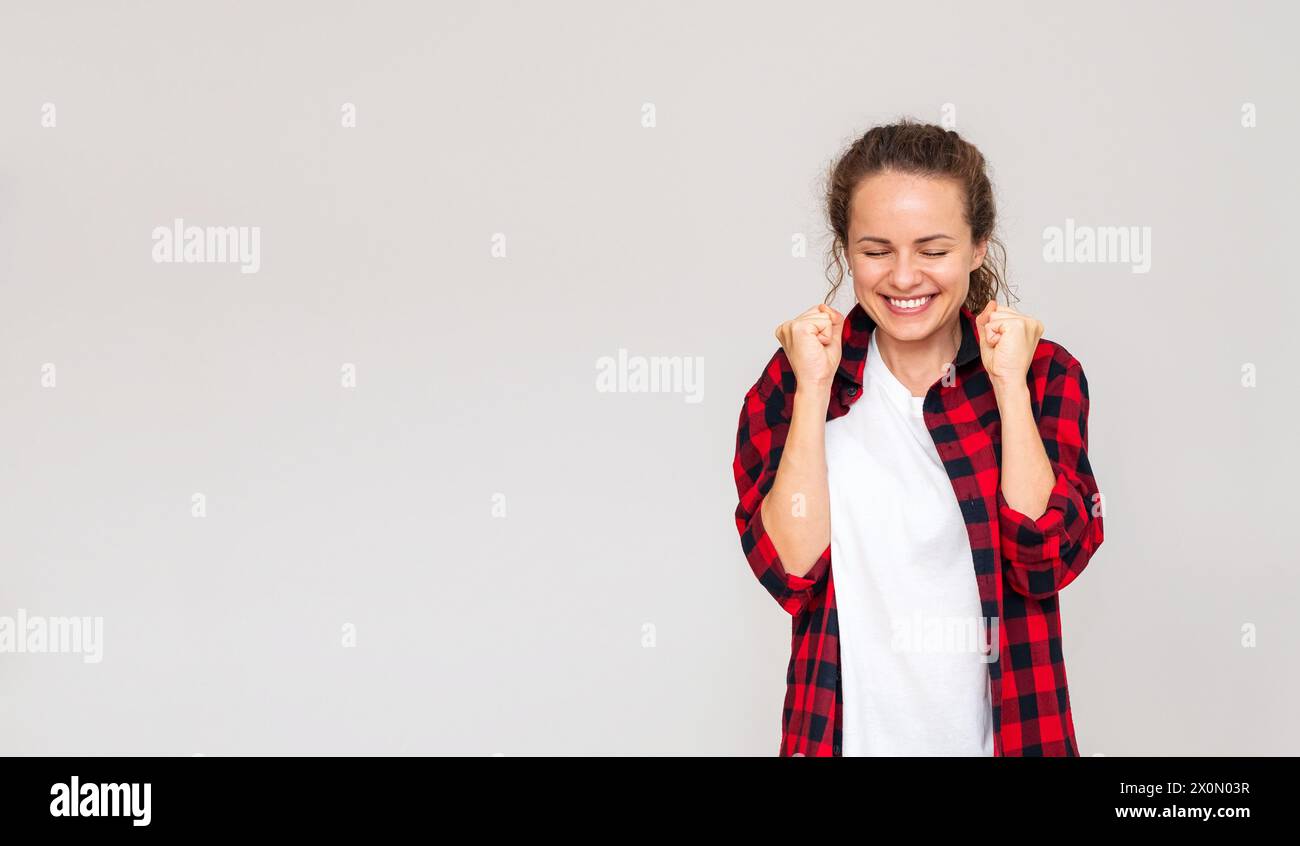Joyful woman. Expression of happiness. A woman rejoices with her eyes closed and her fists clenched on plain background in studio. Stock Photo