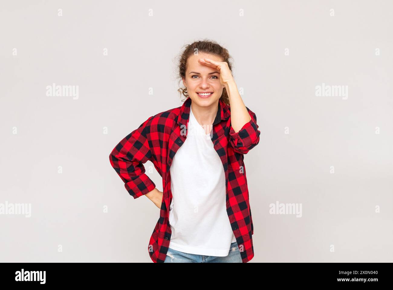 Woman  wearing casual red shirt shading her eyes with hand, looking in to the distance and smiling. Studio shot. Stock Photo