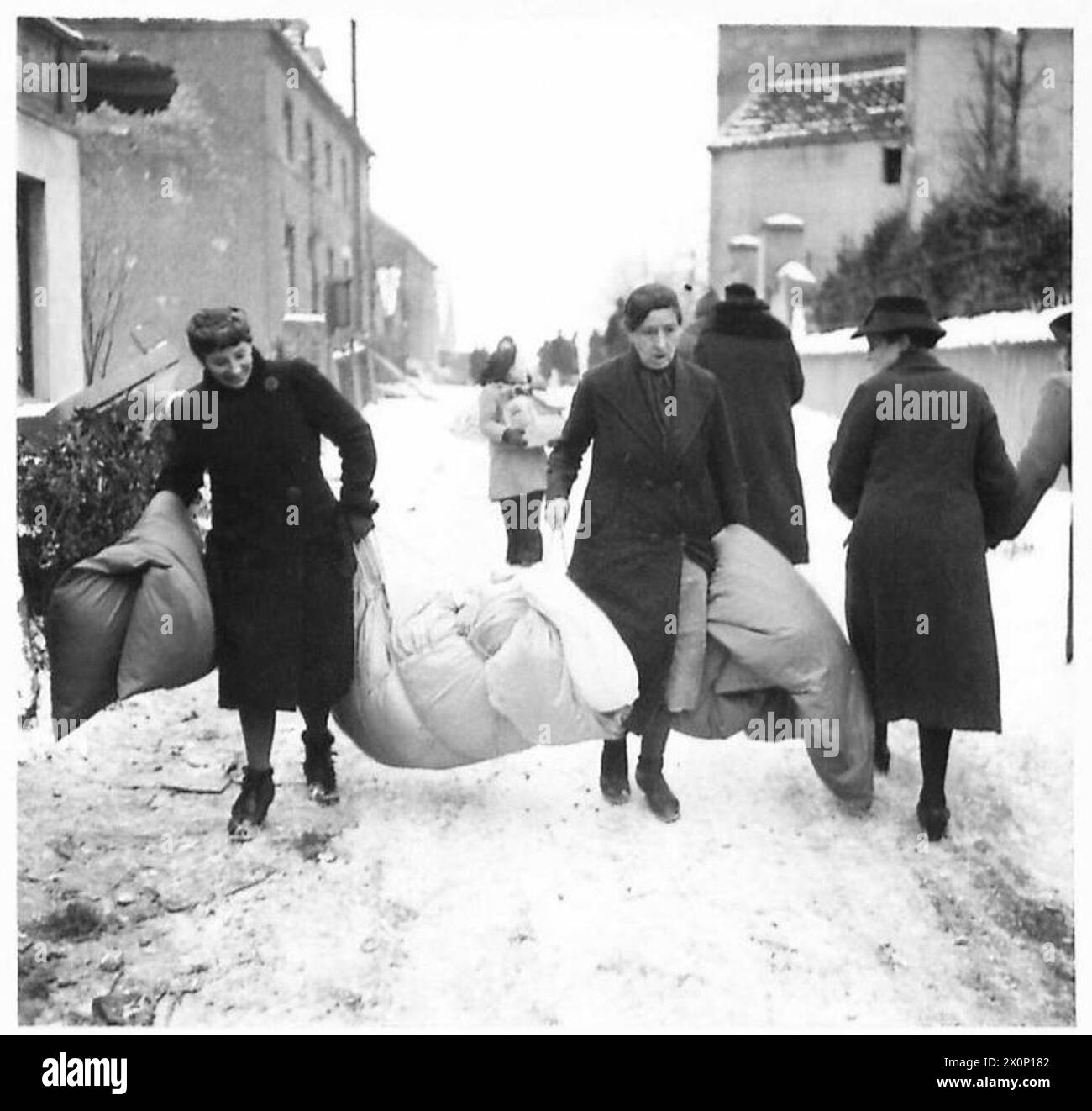 Two German women carry personal belongings to their new home in Tuddern  within the Sittard Triangle. The scene reflects civilian displacement  during World War II in Europe, observed by British Army personnel