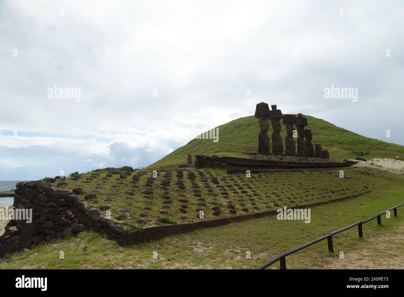 Atop a lush green hill with a dramatic sky overhead, a line of ancient Moai statues oversees the landscape of Easter Island. These iconic figures, car Stock Photo