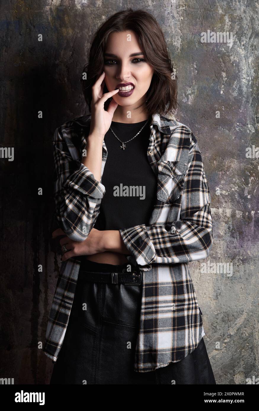Portrait of naughty grunge (rock) girl. Cool informal model, dressed in checkered shirt and leather skirt stands at the wall Stock Photo