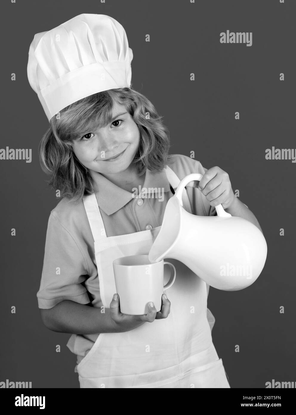 Milk for kids. Child chef cook prepares food in isolated blue studio background. Kids cooking. Teen boy with apron and chef hat preparing a healthy Stock Photo