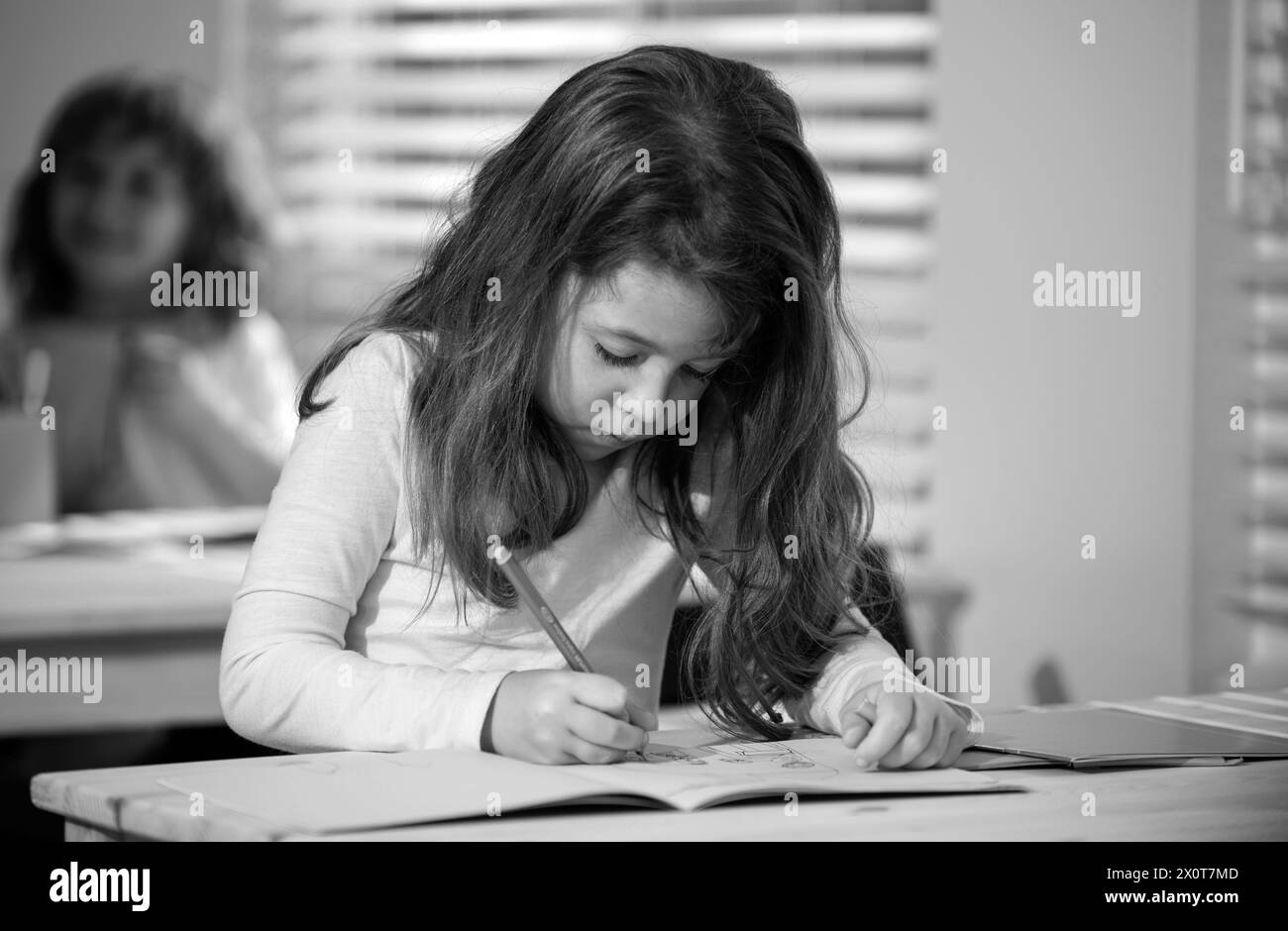 Concentrated schoolgirl sitting at desk and writing in exercise book with classmate sitting behind. Stock Photo