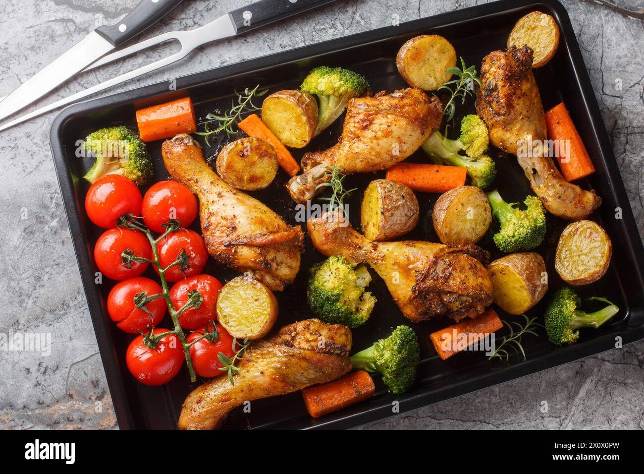 Tasty Baked ruddy chicken legs with potatoes, broccoli, tomato, carrot close-up on a baking sheet on the table. Horizontal top view from above Stock Photo