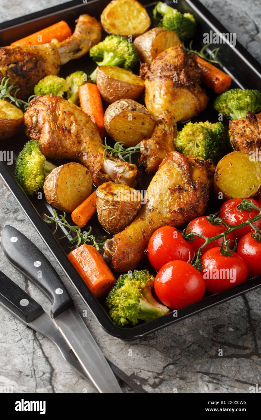 Baked chicken drumstick with broccoli, potatoes, tomatoes, and carrots close-up on a sheet pan on the table. Vertical Stock Photo