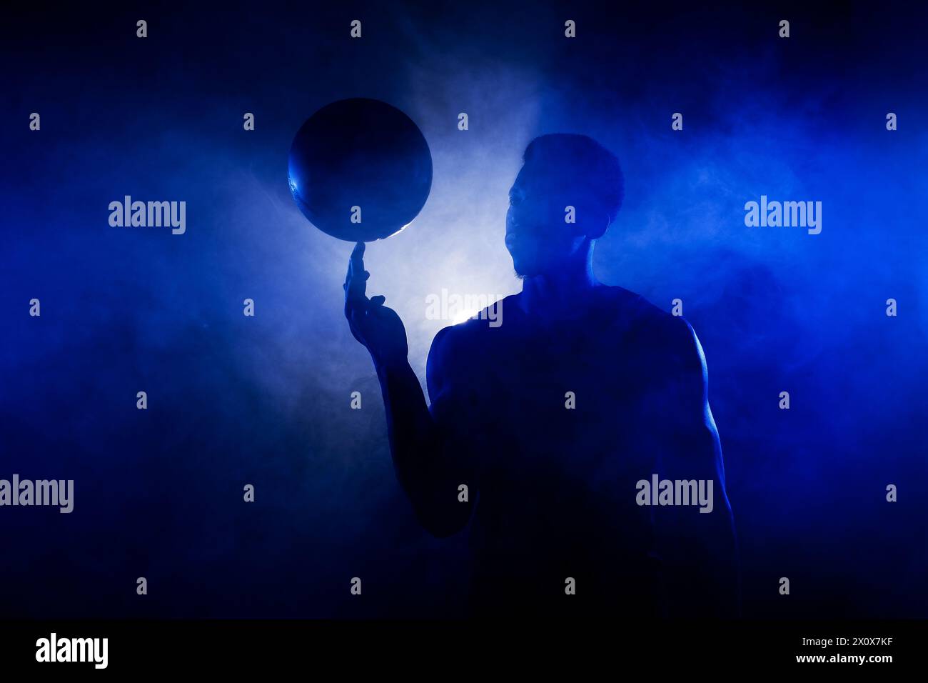 Basketball player silhouette lit with blue color spinning a ball against smoke background. Muscular african american man. Stock Photo