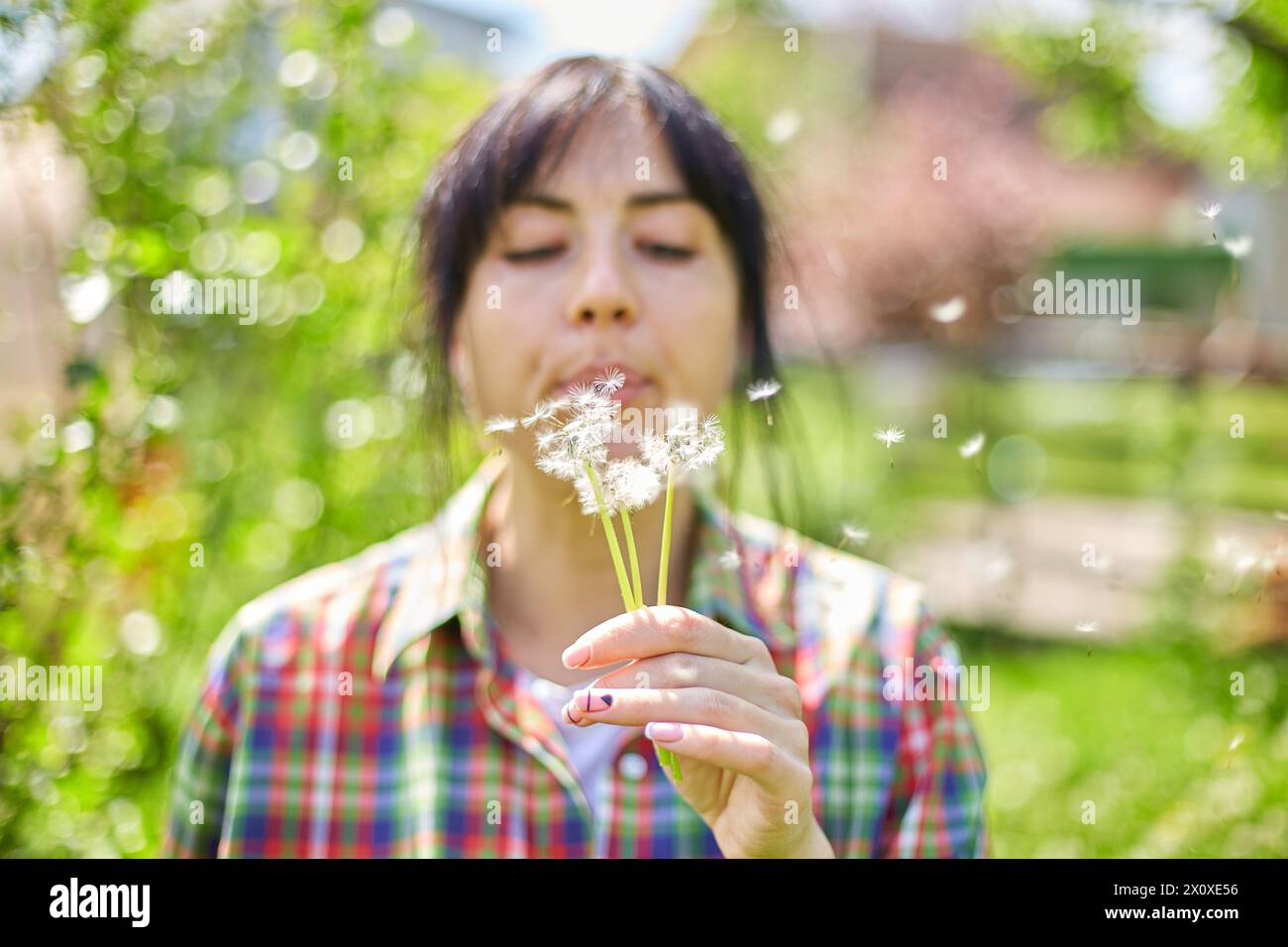 A woman gently blows on a dandelion, scattering seeds in a vibrant garden, Springtime Stock Photo