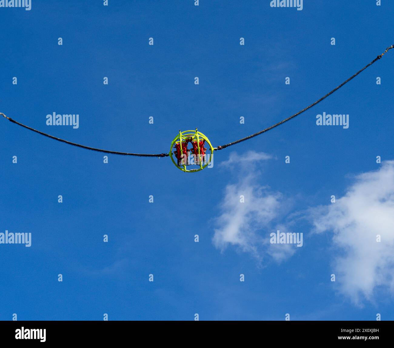 Two thrill-loving friends in a yellow sling soar high into a blue sky with clouds. In a playground during a summer day in Italy. Stock Photo