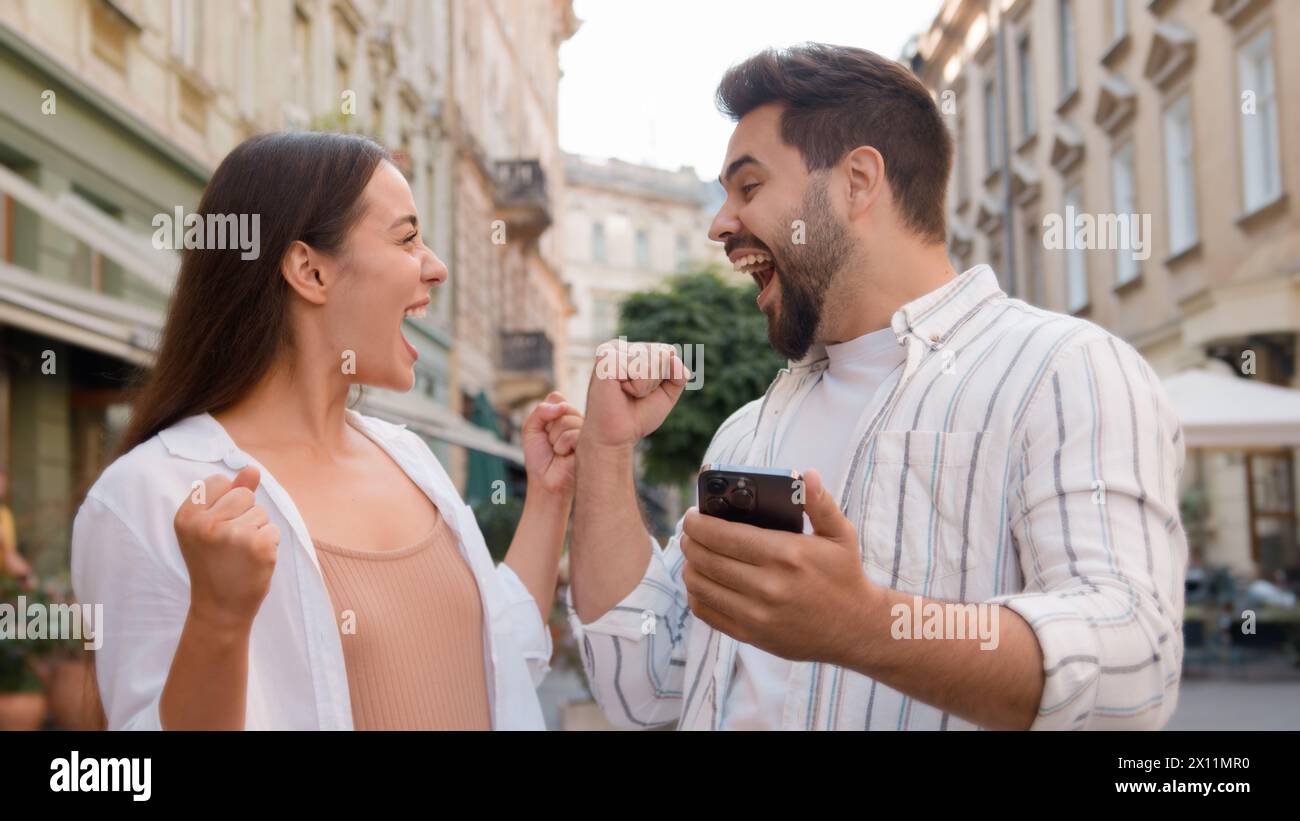 Caucasian couple happy woman man rejoices winning victory lucky expression together joy using mobile phone cellphone high-five exclamation success Stock Photo