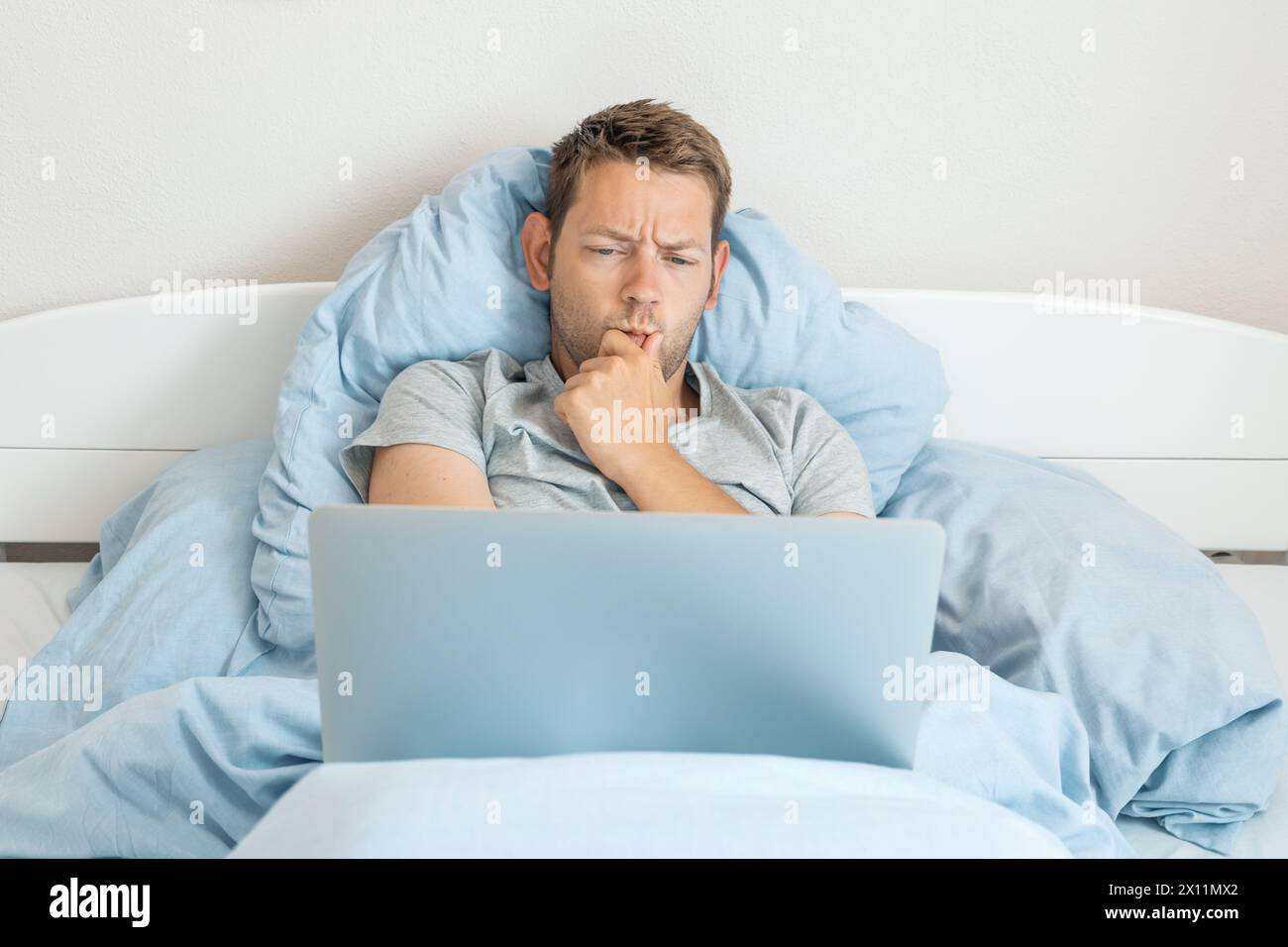 Young thoughtful man lying in the bed and working on a laptop. Serious Caucasian male freelancer surfing and browsing internet, working online from Stock Photo