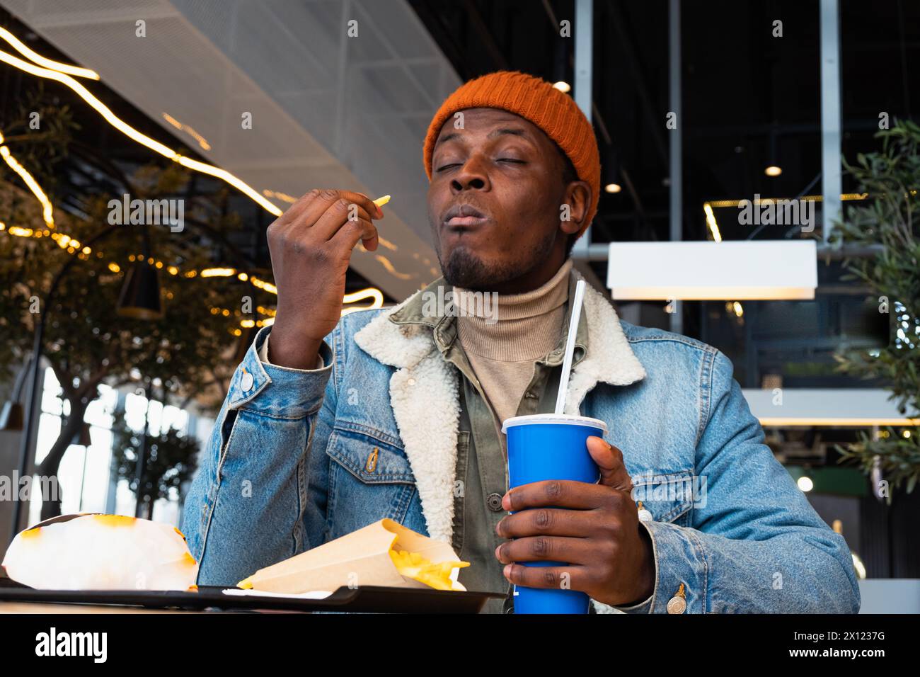 African American man in a denim jacket and orange beanie savors his meal, with a soda cup in hand, in the vibrant ambiance of a contemporary cafe.  Stock Photo