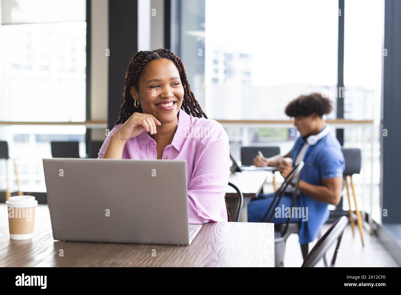 Young biracial woman with braided hair working on laptop, smiling, in a modern business office Stock Photo