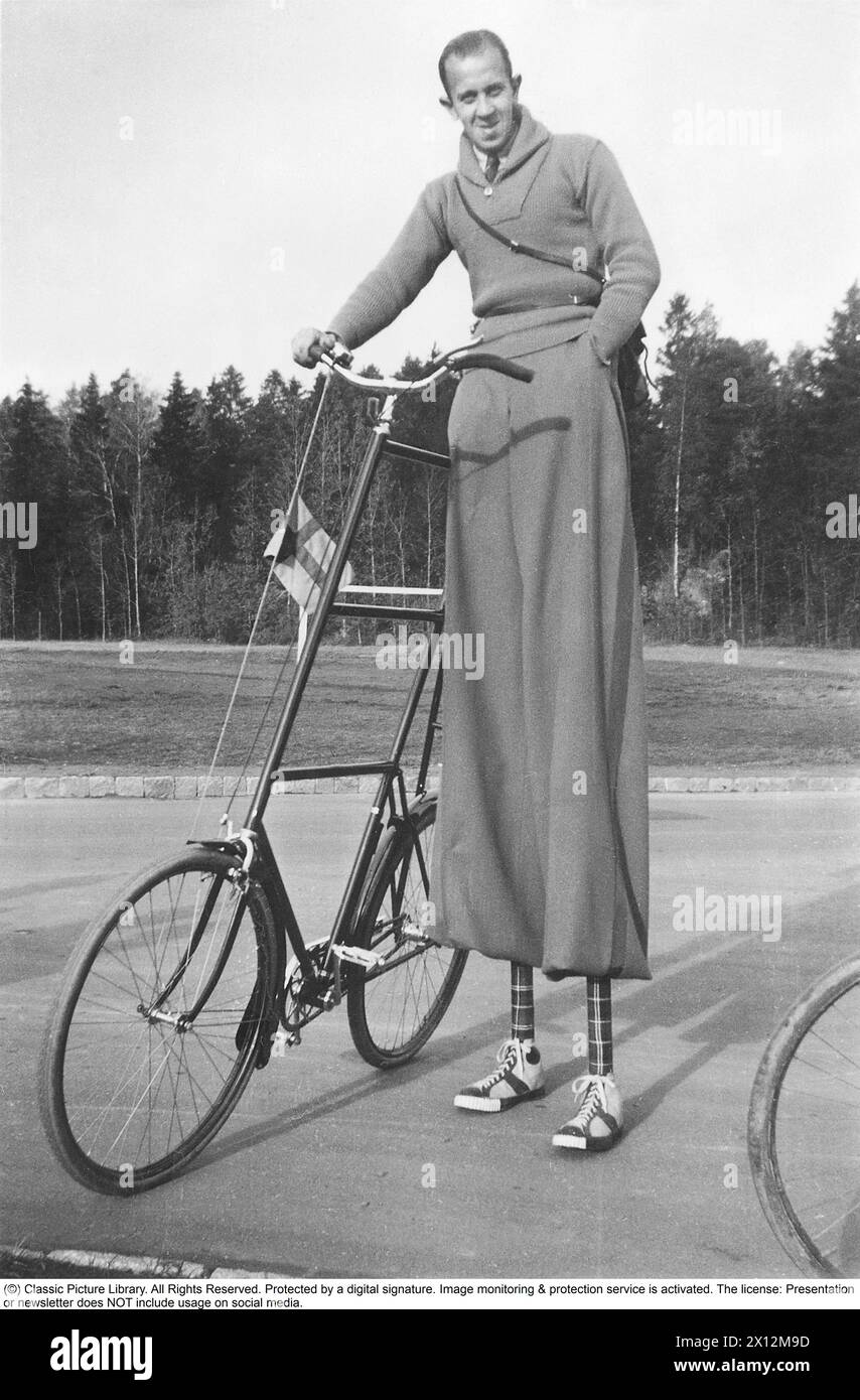 Odd bicycle with equally odd cyclist in the 1940s. A man seen on stilts to be able to ride his taller than usual bicycle. His equally long trousers covers the stilts and they are in the typical 1940s design breeches plus fours. The shoes and the ankles are nicely decorated to complete the overall look of the odd man. 1940 Stock Photo