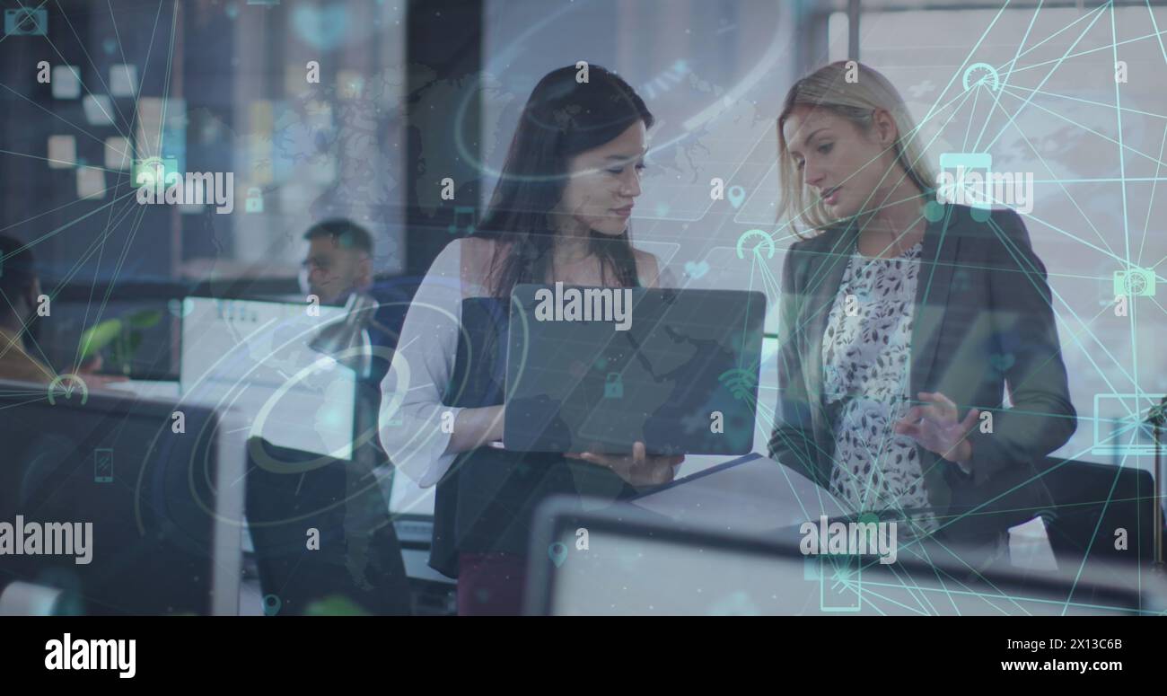 Image of globes of digital icons over two diverse women discussing over a laptop at office Stock Photo