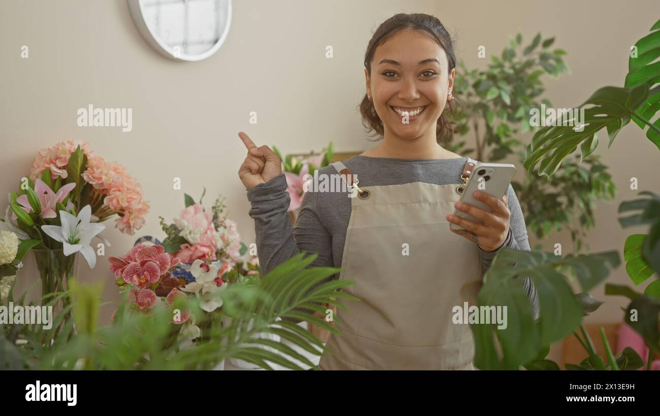Smiling woman in apron holding smartphone and pointing in a flower shop surrounded by plants and floral arrangements Stock Photo