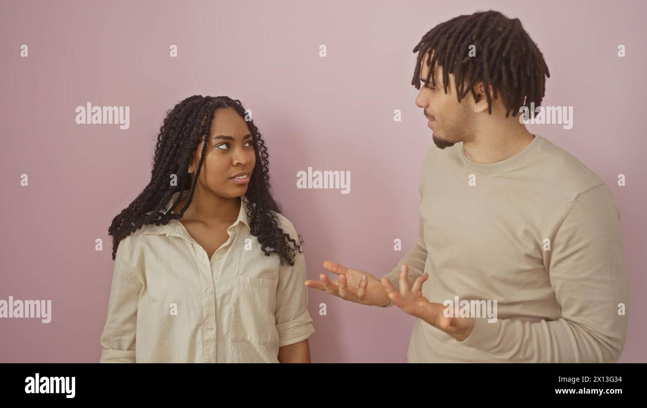 A man and a woman engage in a focused conversation, expressing mixed emotions against a plain pink background. Stock Photo