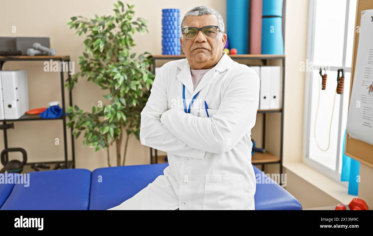 Confident mature man in white lab coat standing in a well-equipped physical therapy clinic room. Stock Photo