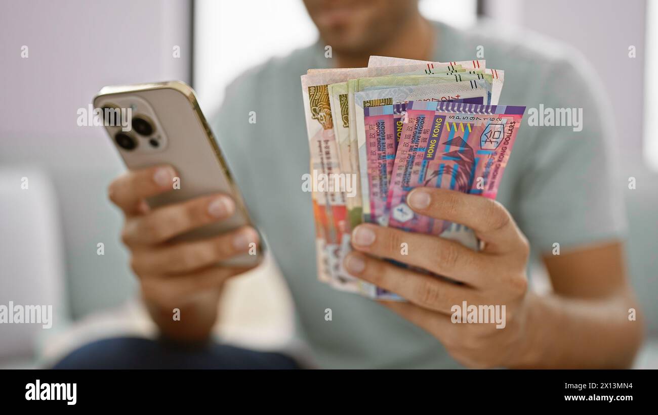 Young man sitting at home on the living room sofa, banking business at his fingertips, holding a stack of crisp hong kong dollar banknotes, glued to h Stock Photo