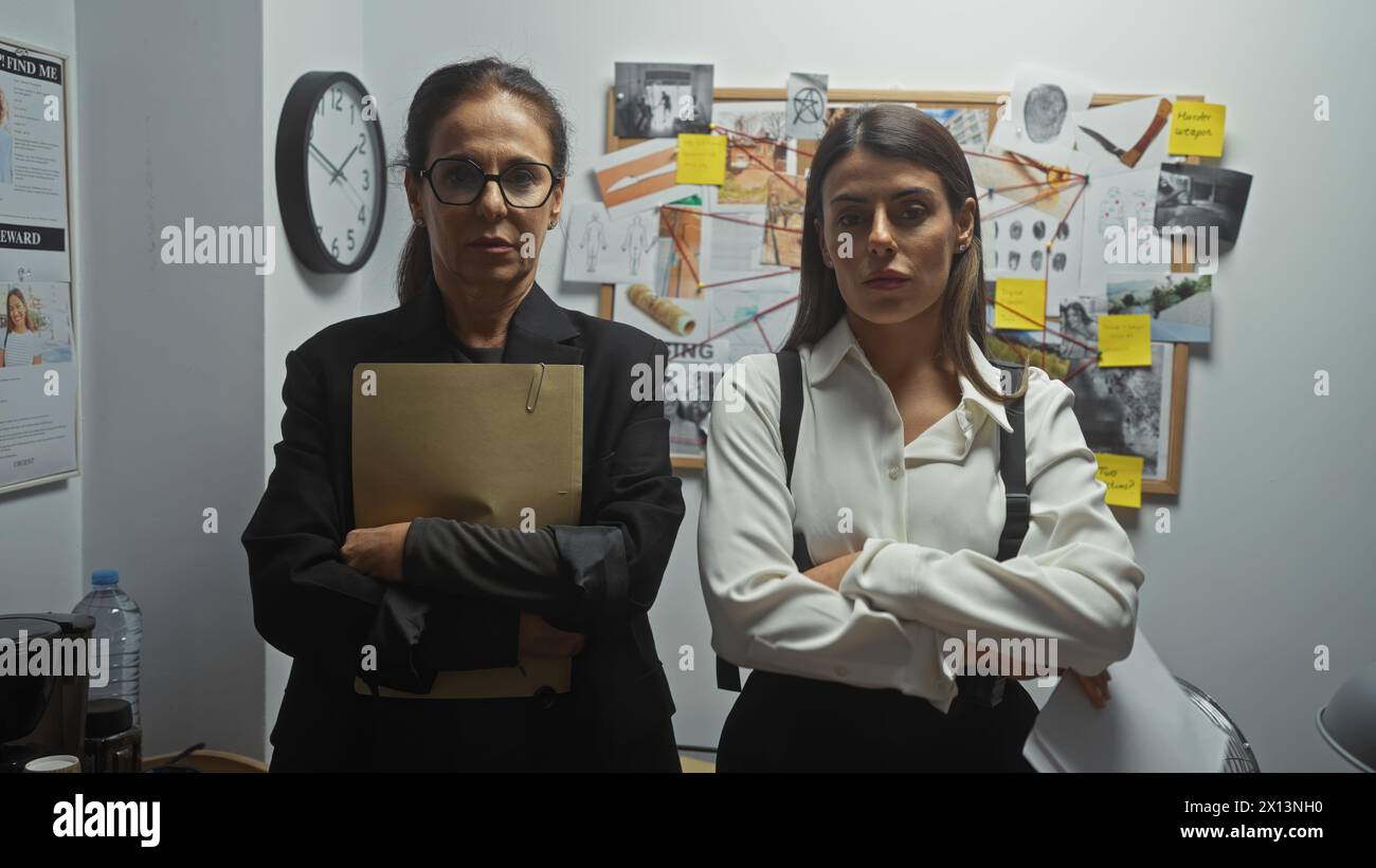 Two serious women detectives stand with crossed arms in a police department investigation room, surrounded by evidence. Stock Photo