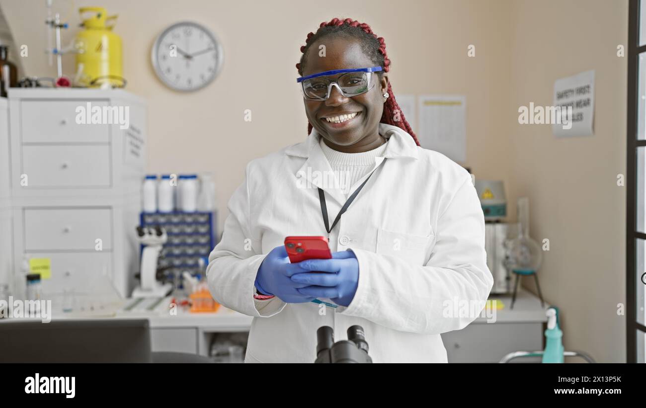 A smiling african american woman in lab coat and gloves using smartphone in a laboratory with equipment and a clock on the wall Stock Photo