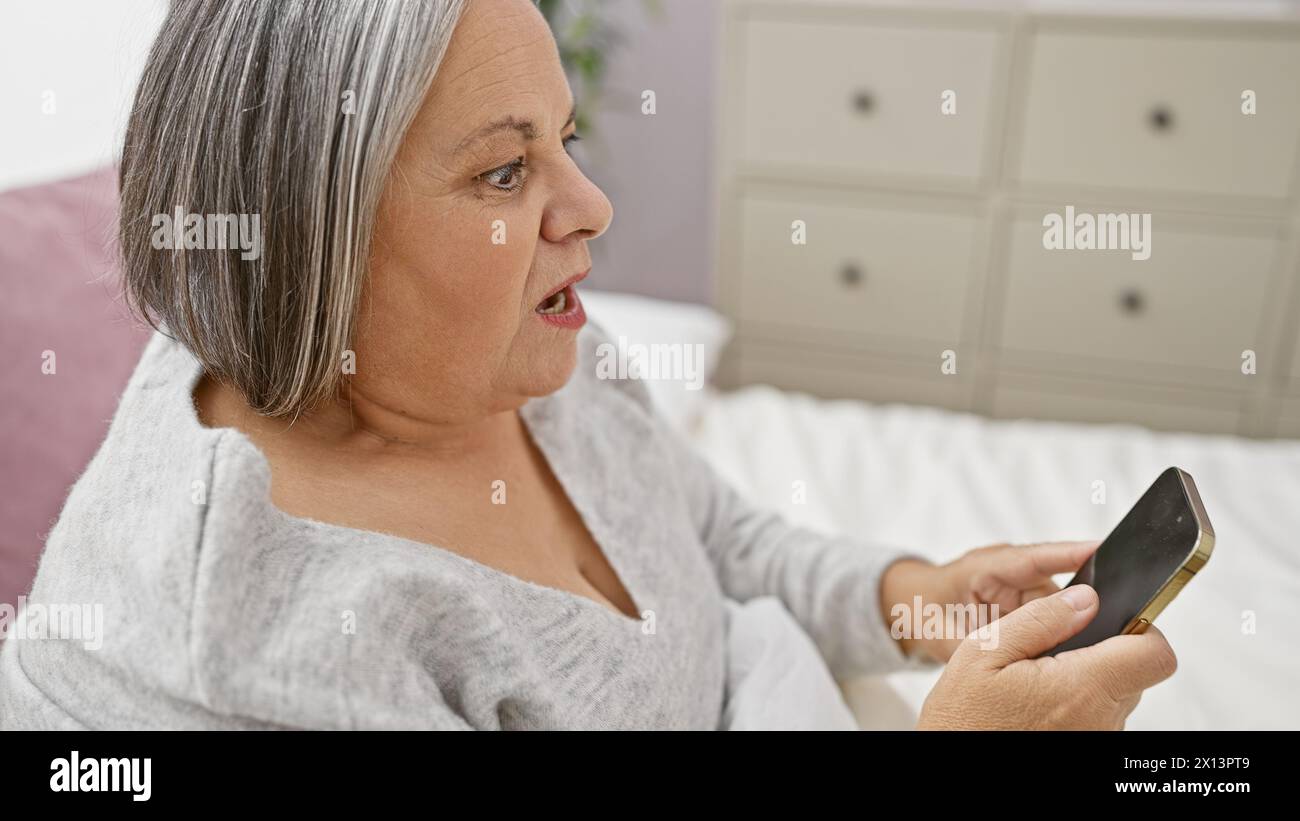 Surprised senior woman with grey hair looking at smartphone in bedroom. Stock Photo