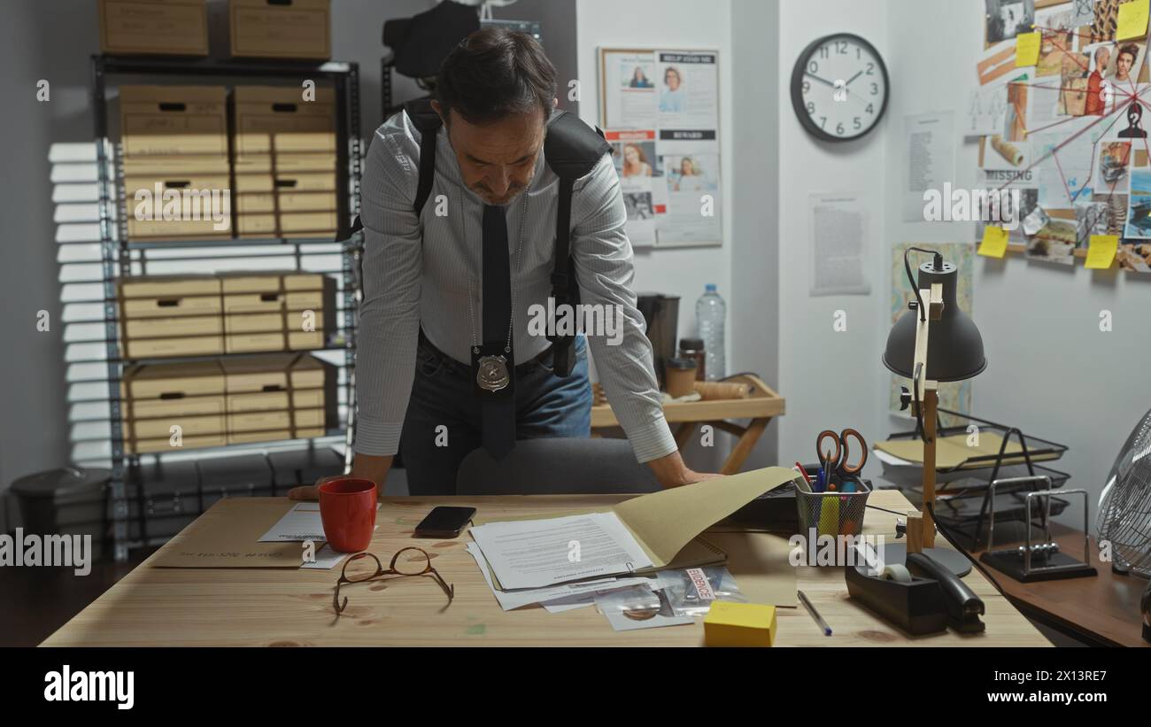 A focused middle-aged detective leans over a cluttered desk analyzing various items in a dimly lit police station office. Stock Photo