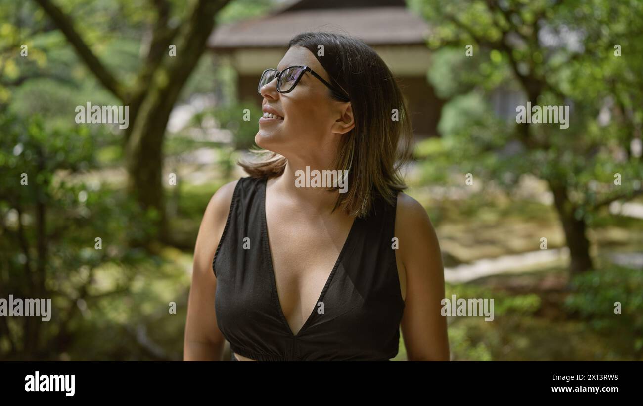 Cheerful young hispanic woman, effortlessly beautiful with glasses, confidently posing and joyfully smiling while exploring the serene surroundings of Stock Photo