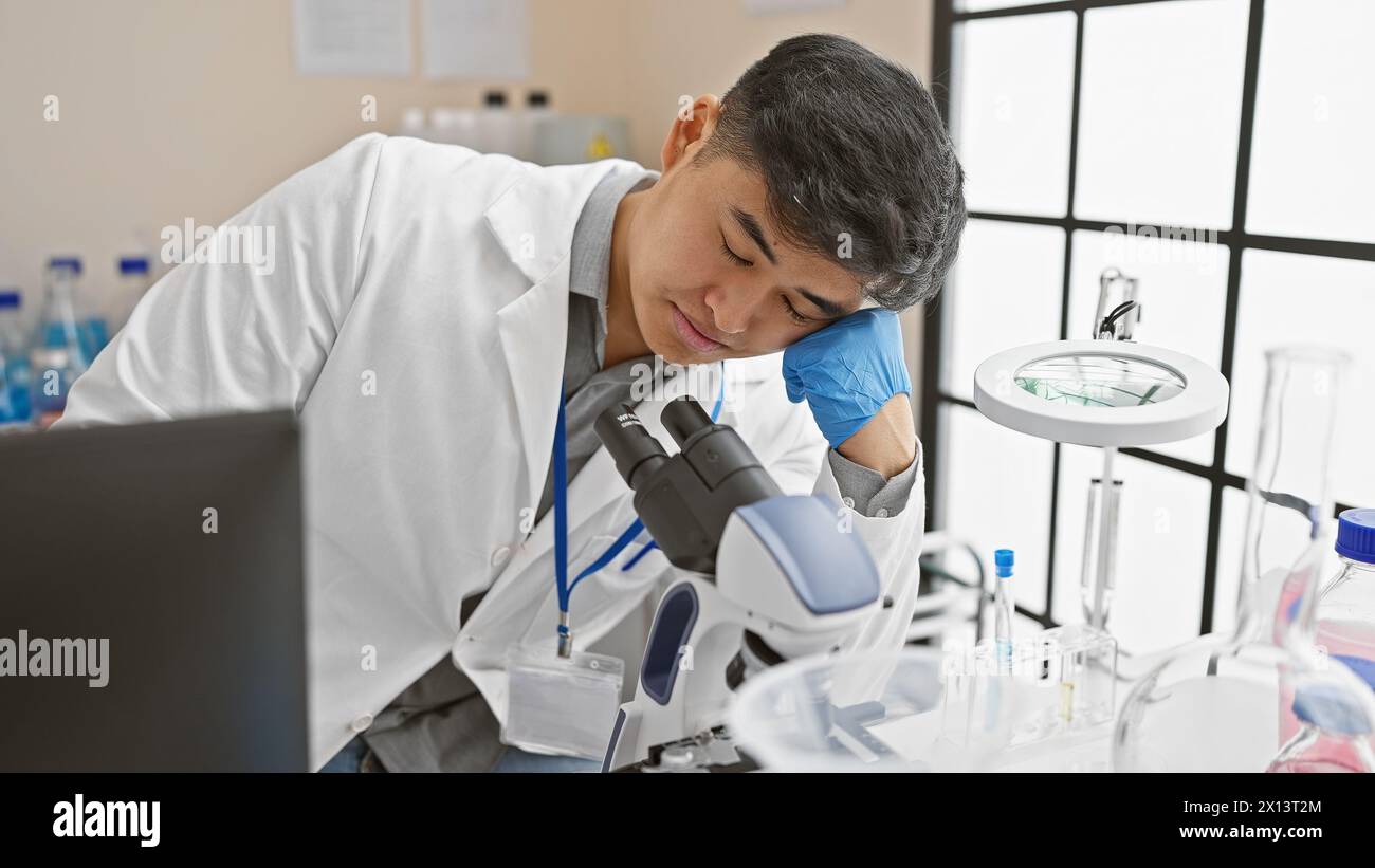 A young asian man in a lab coat appears exhausted while working with a microscope in a laboratory setting. Stock Photo