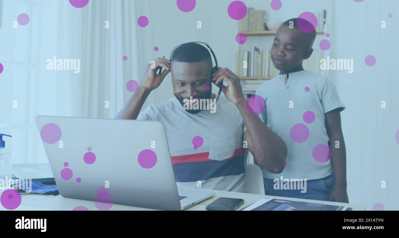 Image of pink spots floating over african american father and son using phone headset and laptop Stock Photo
