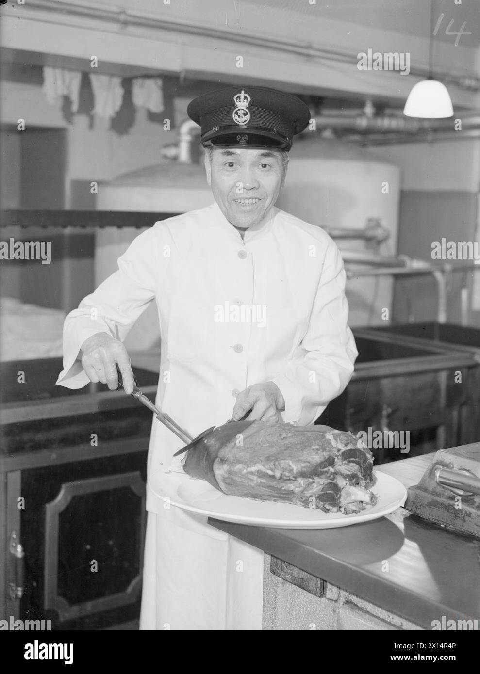 SERVED IN 29 SHIPS. 16 MAY 1944, HMS DINOSAUR, COMBINED OPERATIONS BASE, TROON. 65 YEAR OLD PETTY OFFICER COOK SEI TAN, OF HONG KONG, NOW CHIEF COOK IN THE KITCHEN AT HMS DINOSAUR, AND IN CHARGE OF 22 OTHER COOKS. HE JOINED THE NAVY IN 1904 AND HAS SERVED IN 29 SHIPS. - Petty Officer Cook Sei Tan carving a ham in the galley Stock Photo