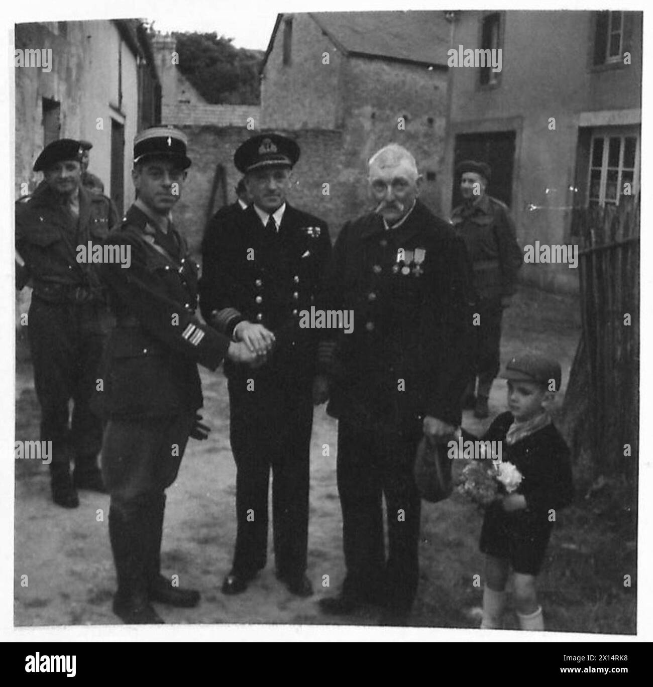 14 JULY CELEBRATIONS AT ARROMANCHES - A French Liaison Officer, Colonel Lion, and Captain H. Hickling, DSO, RN, cordially greet an old French soldier M. Leon Descouvreur, who was a sergeant in the last war, and fought at Verdun. With him is his little grandson whose father is in German hands. Medals:- Victory Medal; Croix des Ancients Combatants, Croix d'Union des Ancients Combatants, Verdun Medal British Army, 21st Army Group Stock Photo