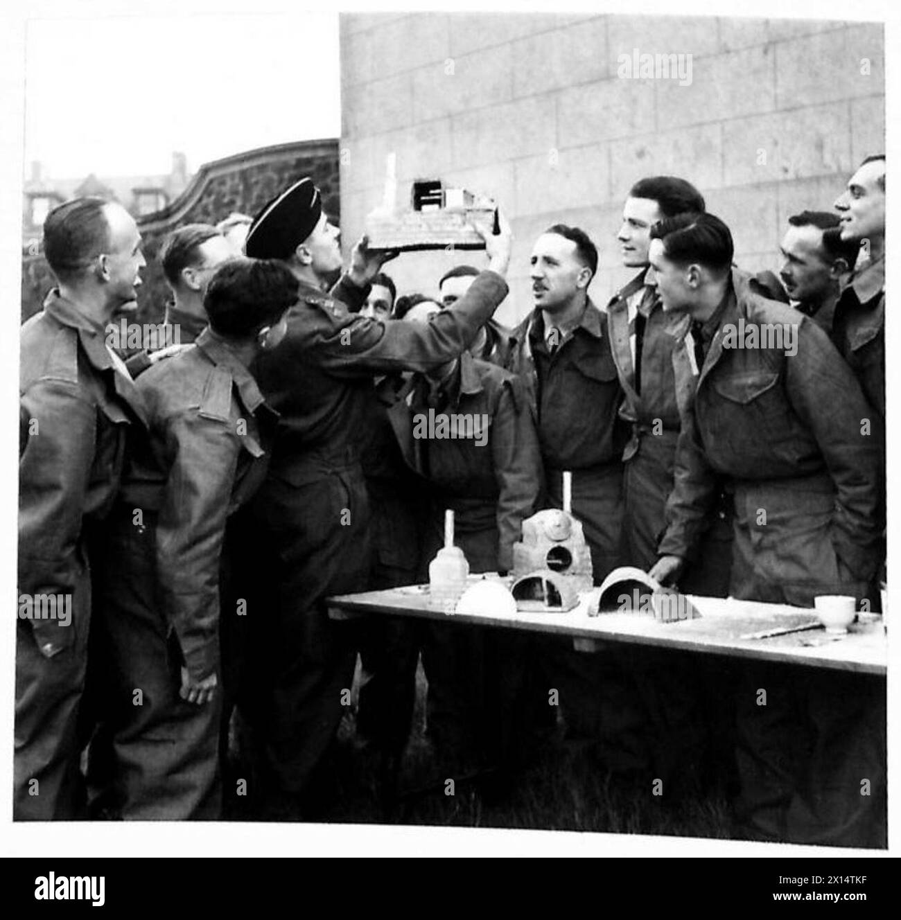 MESSING OFFICERS LEARN TO COOK - An improved oil and water fire made up by using two bully beef tins into a stove, is demonstrated by a visiting catering adviser. The fuel tank is a treacle tin and the oil (waste oil from sumps etc) is fed by trickling from a hole in the fuel tin down a channel of tin. Control of feed is by means of a nail in the hole of the tin. One of the students takes his turn before his fellows as 'pastry cook'. It is significant that the officer students actually prefer to eat their own preparations to those of the regular cooks British Army Stock Photo