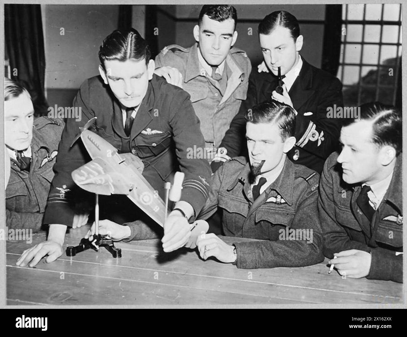 EMPIRE CENTRAL FLYING SCHOOL : AIR UNIVERSITY OF THEALLIED NATIONS. - (Picture issued 1943) 8186. Officers at the E.C.F.S. studying how air flows past an aircraft in flight. A wind tunnel mounted on the bench provides an air stream over the model aircraft, which can be tilted to various flight altitudes. The flow over the wings is demonstrated by the behaviour of the tufts of wool on their upper surfaces. The wing-tip air-vortices are shown by the spinning of the small windmill held by the Australian wing commander Royal Air Force Stock Photo