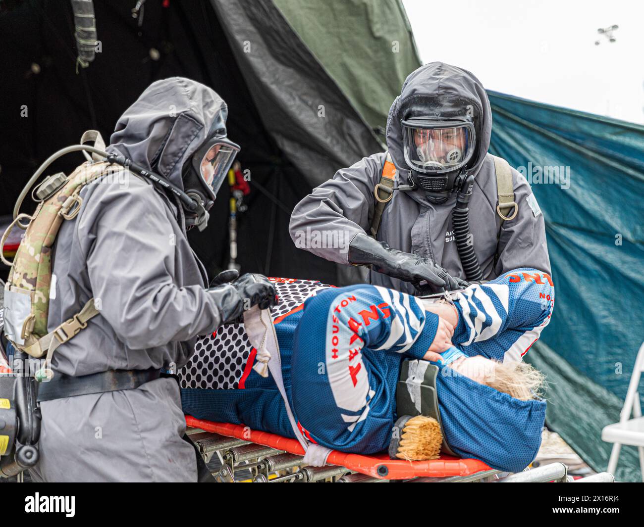 Two people in hazmat suits are tending to a person on a stretcher. The person on the stretcher is wearing a blue and white shirt. Stock Photo