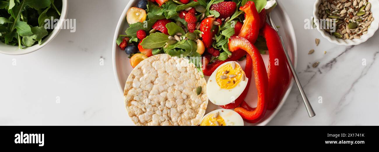 Healthy tasty breakfasts banner, a plate with boiled eggs and rice bread, salad of arugula and strawberries with cherries and blueberries, fresh peppe Stock Photo