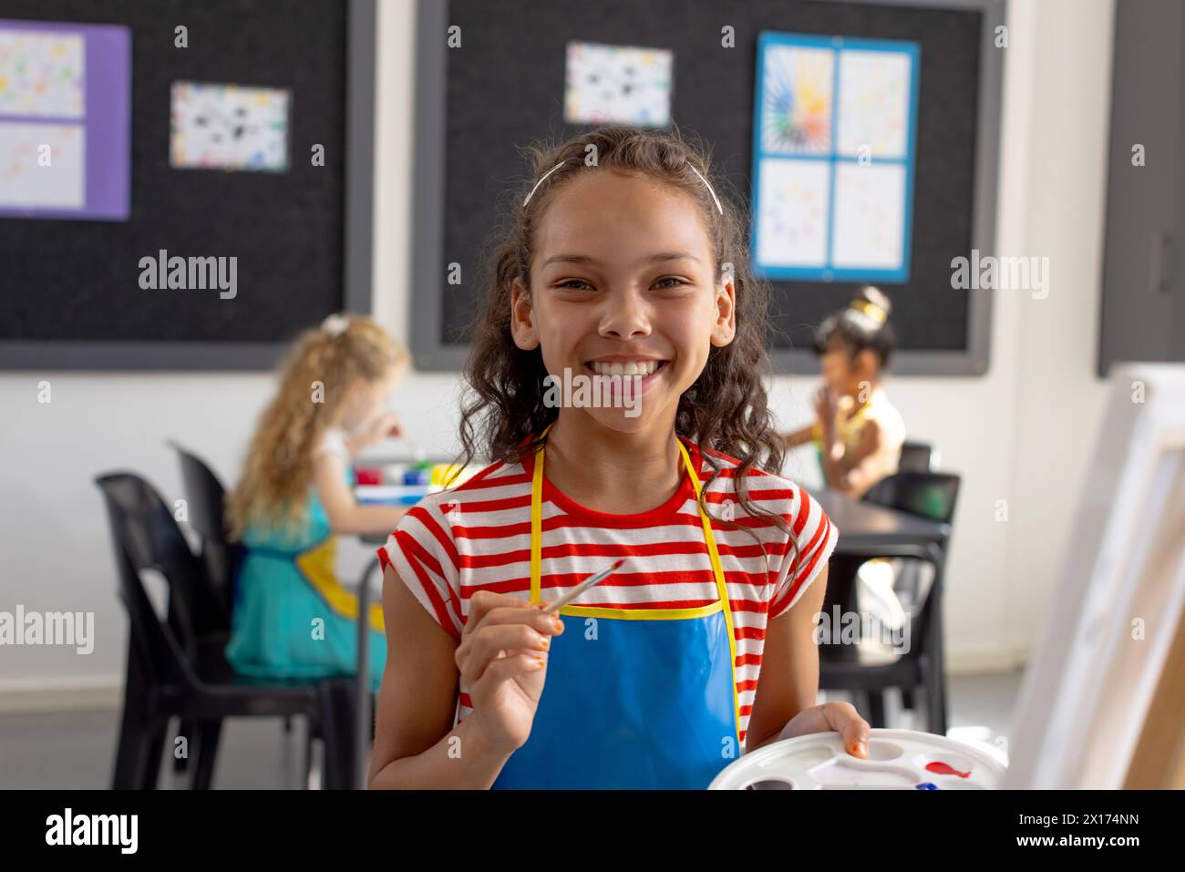 In school art class, biracial young girl holding a paintbrush smiles at camera Stock Photo