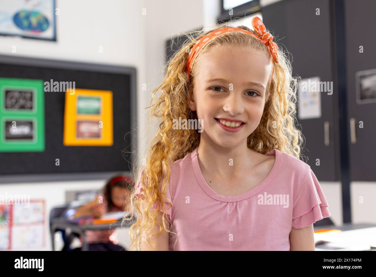 In school, young Caucasian girl with curly blonde hair is smiling in a classroom Stock Photo