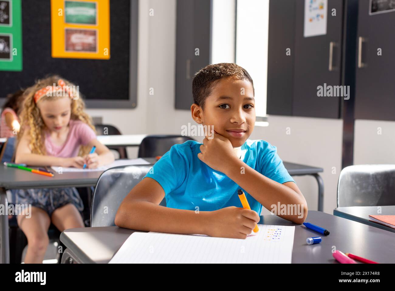 In school, young biracial male student sitting at a desk in a classroom, looking thoughtful. Behind him, a biracial girl and a younger Caucasian girl Stock Photo