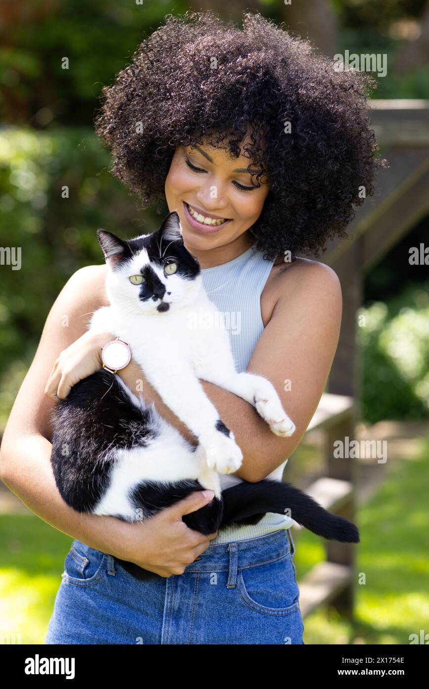Biracial young woman holding black and white cat, both outdoors at home in garden Stock Photo