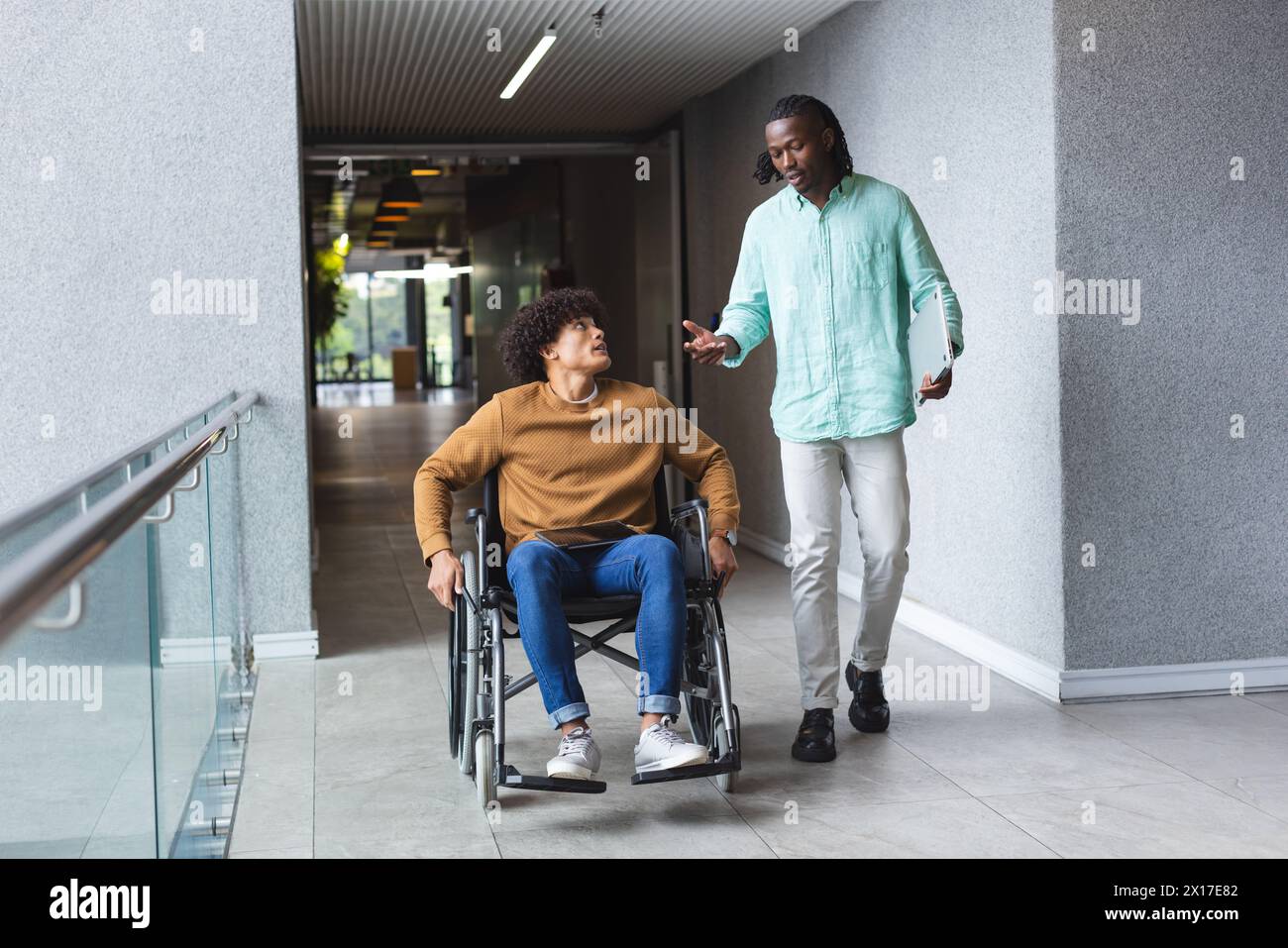 African American man pushing wheelchair of biracial man, both talking in a modern business office Stock Photo