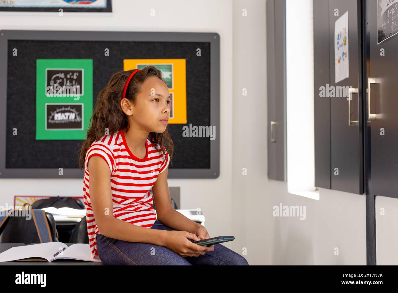 In school, in classroom, young biracial girl holding a smartphone looks thoughtful Stock Photo