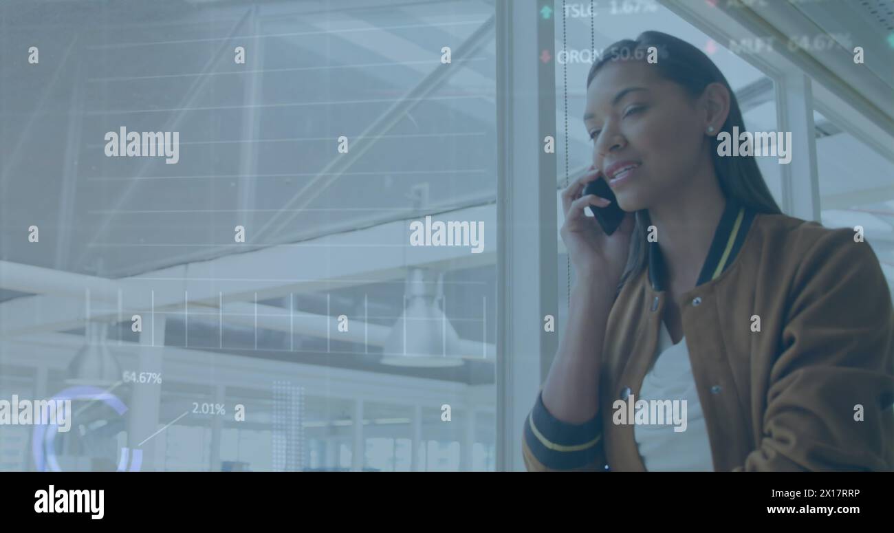 Image of graphs, circles and trading board, biracial businesswoman talking on phone in corridor Stock Photo
