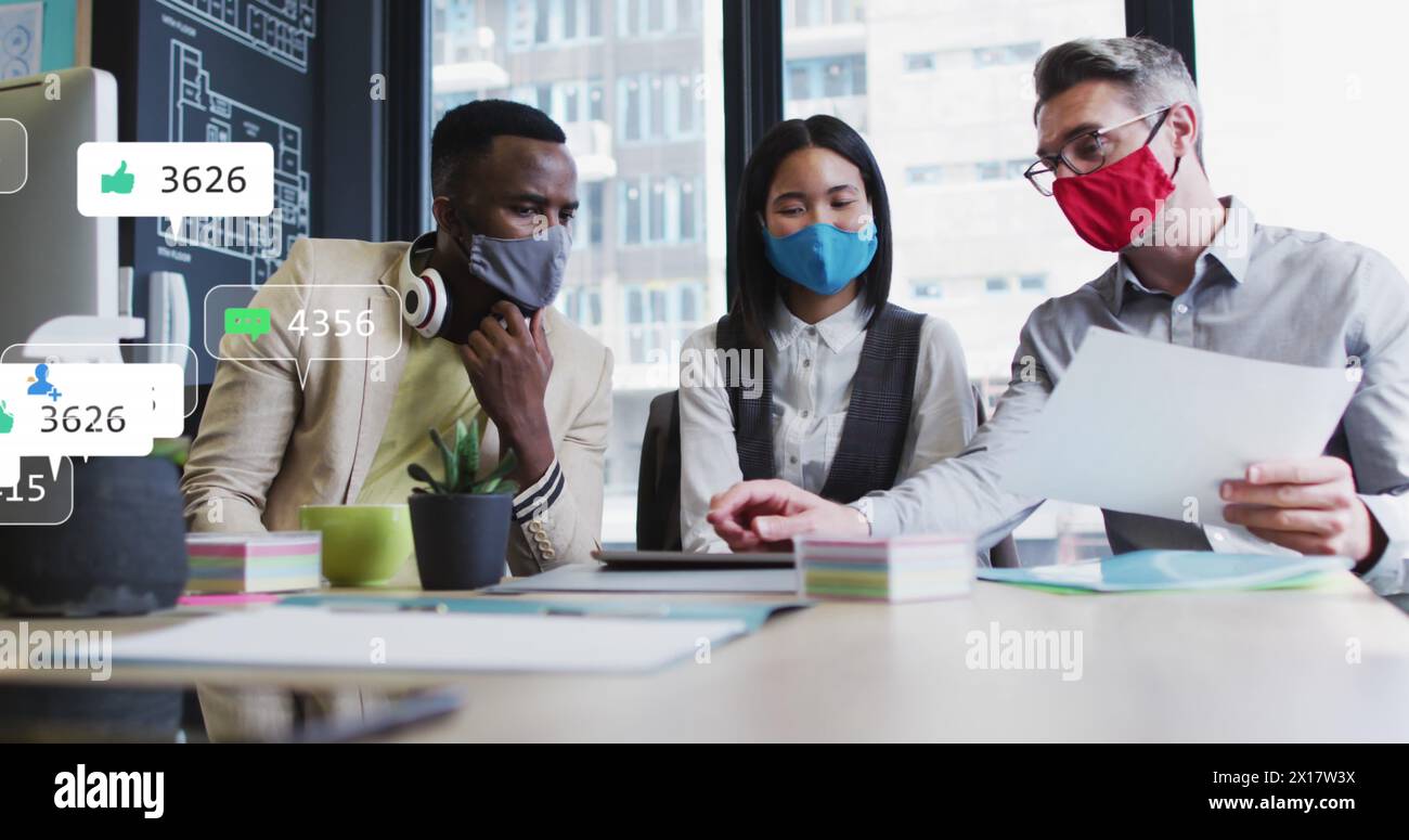Image of social media icons over diverse colleagues wearing face masks discussing at office Stock Photo
