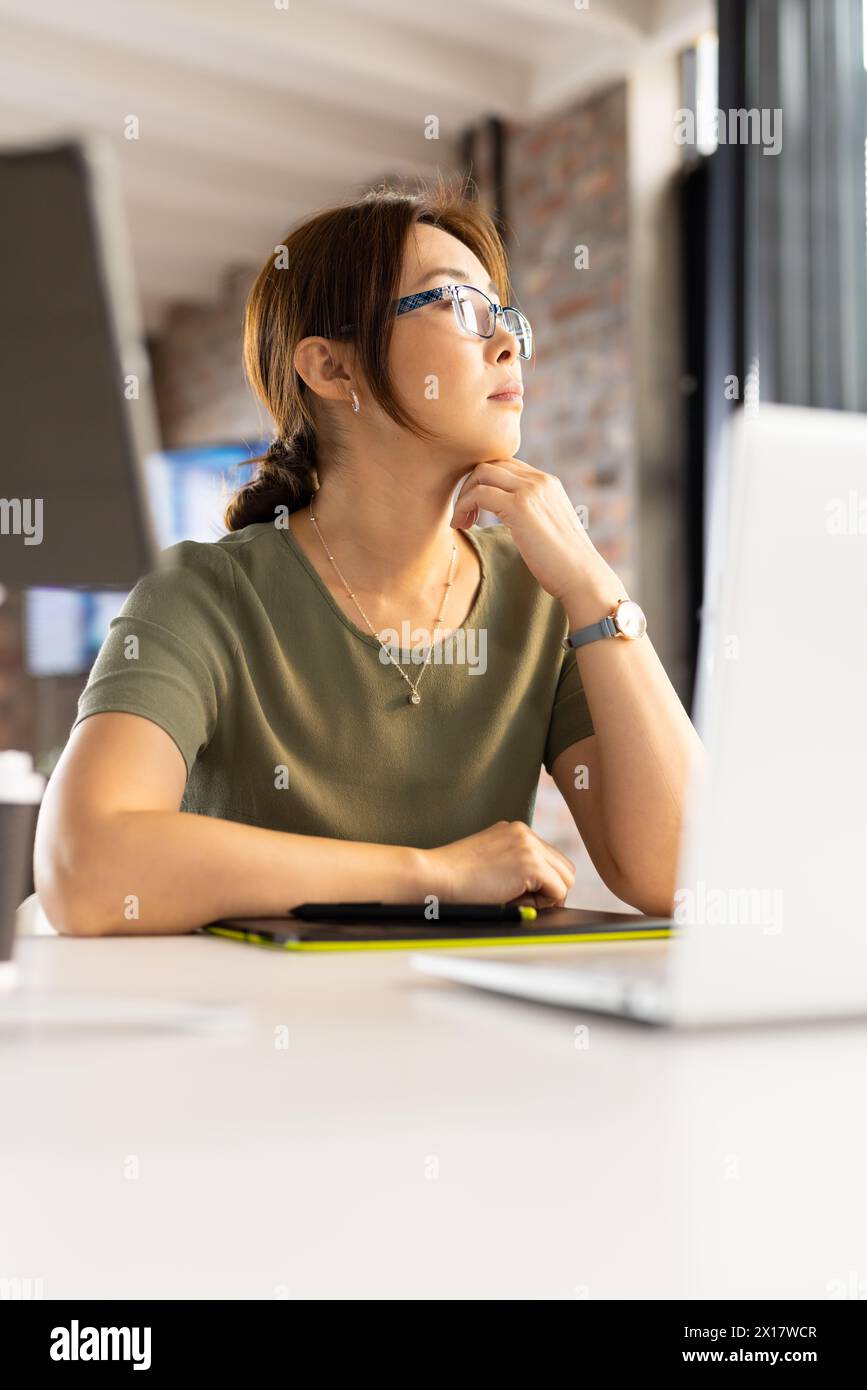 Middle-aged Asian woman wearing glasses looking at laptop screen in a modern business office Stock Photo