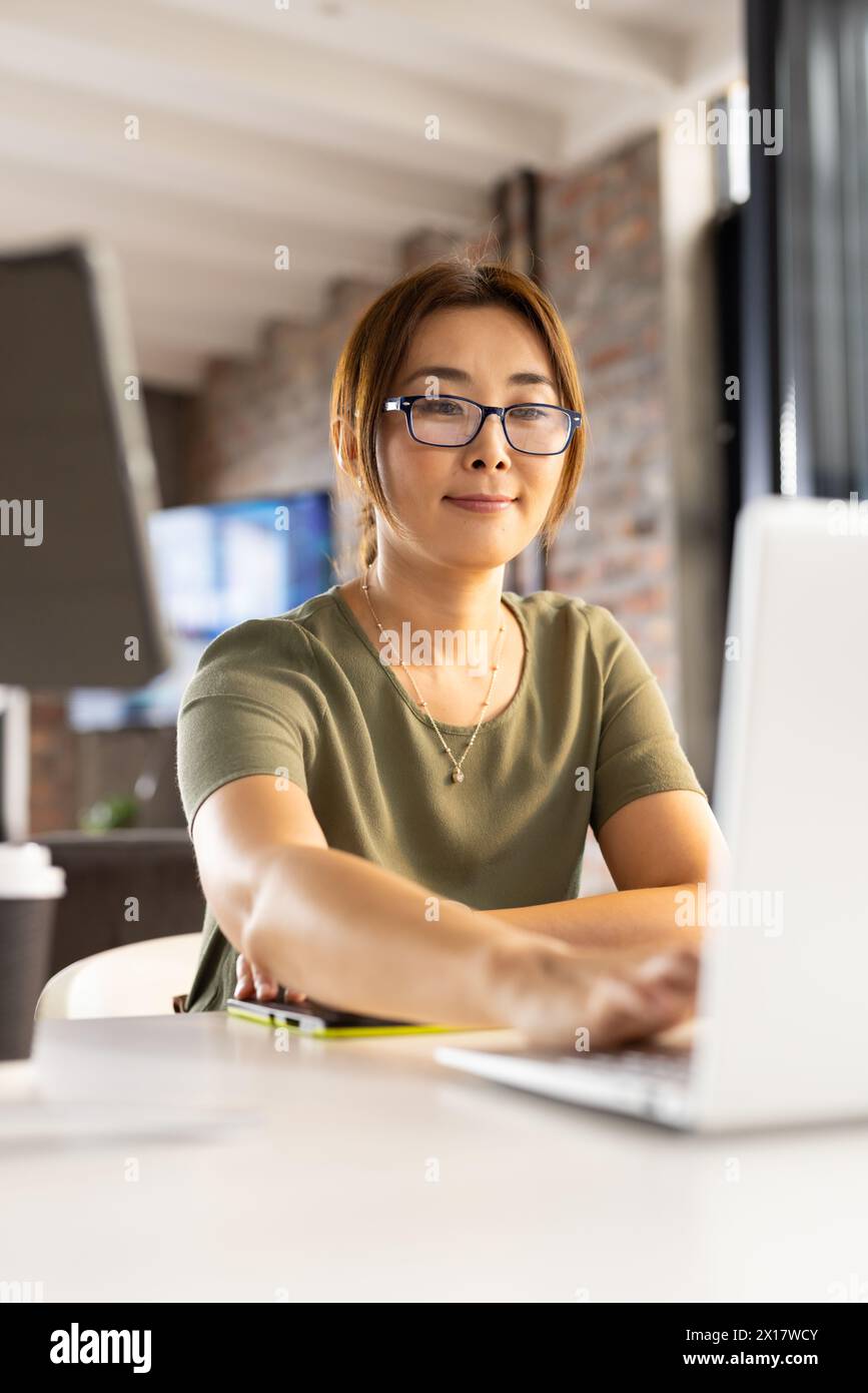 Middle-aged Asian woman wearing glasses, working on a laptop in a modern business office Stock Photo