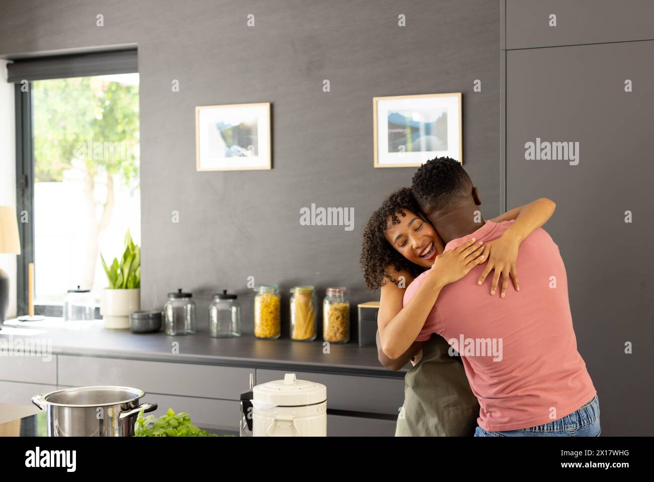 Diverse couple embracing in kitchen, near wall art with copy space Stock Photo