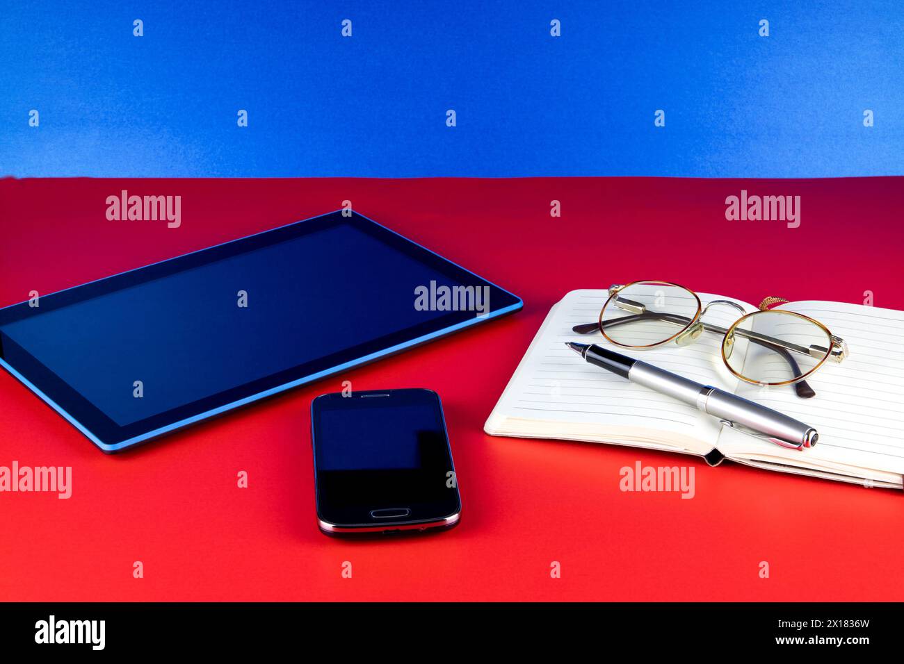Mobile phone with tablet device and notebook pen and eye glasses on a red base and blue background Stock Photo
